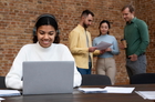 Smiling woman in headphones working on a laptop with colleagues discussing documents behind her.